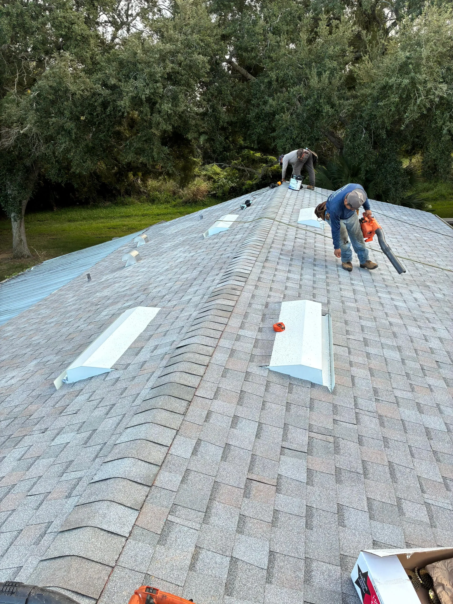 H&H Roofing workers on a sloped shingled roof performing maintenance with tools and a toolbox nearby, green trees in the background repeatedly visible behind them.
